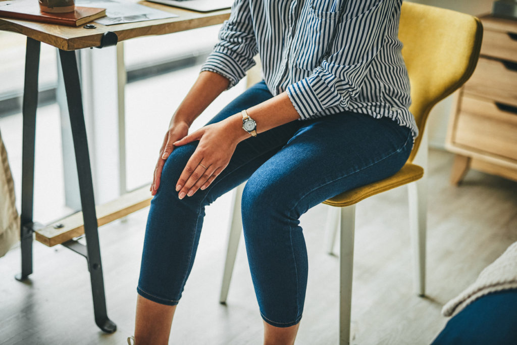 Woman in blue jeans sitting in a chair and holding her knee demonstrating pain coming from the area.