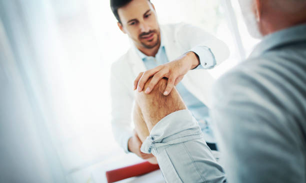 Closeup over the shoulder view of an early 60's senior gentleman having some knee pain. He's at doctor's office having medical examination by a male doctor. The doctor is touching the sensitive area and trying to determine the cause of pain.