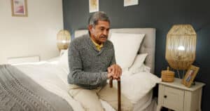 A senior man with osteoporosis is holding a walking stick and reflecting in his bedroom.